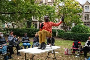 A ZCMP participant with an orange sweater, gold pants, and black shoes sits on a chair on top of a table on a lawn, smiling to his left with a raised gloved hand, with amuse participants staring up, as part of a living image exercise during a workshop.