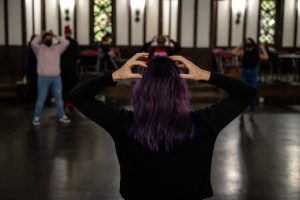211106_MariaJoseWSGroup1_001 ZCMP workshop leader María José, their back towards the camera, participates in the workshop. They hold their hands up to their head, only their fingers grazing their purple hair, as other participants in the background imitate the gesture.