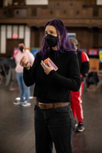 211106_MariaJoseWSGroup1_005 ZCMP workshop leader María José, with their purple shoulder-length hair, black long-sleeve sweater and jeans, and brown belt holds up a lit match in their right hand and the box close to their chest in the other. They convey a longing expression to the person hearing their story out of frame.