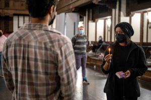211106_MariaJoseWSGroup1_006 Two ZCMP participants face each other in a dimly lit church space. The participant to the right, in dark clothes and beanie, holds up a lit match in their right hand as they look intently at the other participant in a red-blue-gray plaid shirt.