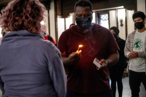 211106_MariaJoseWSGroup1_009 Z huddled group of ZCMP participants in a dimly lit church space. One participants in a red tee shirt faces another participant with pink hair and lavender sweater; the former holds up a lit match as they stare into the fire, a puff of smoke over their face, while the latter waits.