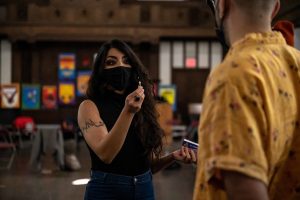 211106_MariaJoseWSGroup1_016 Two ZCMP participants face each other in a dimly lit church space. The participant facing the camera, with long wavy dark hair, black sleeveless top, and blue jeans holds up a lit match in their right hand as they speak.