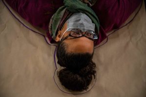 211106_MariaJoseWSGroup1_019 A ZCMP with glasses, hair in a high bun, and green scarf lay on their back over a large rectangle of brown paper as their silhouette is being traced with a purple marker.