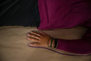 211106_MariaJoseWSGroup1_020 A ZCMP wearing a silver ring, various bangles, black pants and dark red-purple hoodie lay on top of a large brown rectangle of paper as their silhouette is being traced with a purple marker.