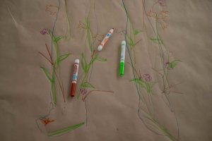 211106_MariaJoseWSGroup1_025 A close-up of a life-sized body map's feet traced in blue lines and decorated with a variety of weeds and lavender flowers. A brown, light green and uncapped orange marker lay over the brown paper figure.