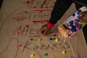 211106_MariaJoseWSGroup1_026 A close-up of a ZCMP participant's hands as they decorate their body map with colorful pompoms. The words FEAR, TIGHT, HELPLESS, and KINDNESS in thick black script are surrounded by red squiggly lines.