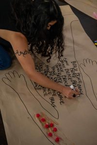 211106_MariaJoseWSGroup1_029 A ZCMP participant with wavy black hair covering their face hunches over their life-sized body map which they have decorated with glittered glue, red pompoms and the word BRACE in thick black script being repeated across the body map's lower half.