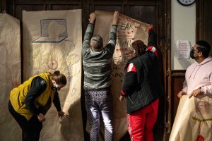 211106_MariaJoseWSGroup1_030 Four ZCMP help hang up colorful life-sized body maps on the back wall of a dimly lit church space.