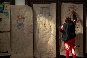 211106_MariaJoseWSGroup1_031 A participant with a red tracksuit and black vest works one on of the life-sized body maps that hang on the back wall of a dimly lit church space. They write the word FEAR in the space of the body's head surrounded by red squiggly lines and pompoms.
