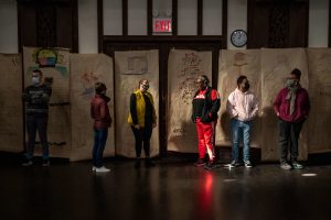 211106_MariaJoseWSGroup1_035 Seven ZCMP participants stand in a dimly lit church space in front of a row of colorful life-sized body maps that hang in the back wall. Most of them look towards different directions, hands in their jacket pockets.