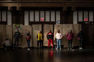 211106_MariaJoseWSGroup1_036 Seven ZCMP participants stand in a dimly lit church space in front of a row of colorful life-sized body maps that hang in the back wall. Most of them look towards the left-hand side, hands in their jacket pockets.