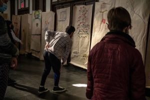 211106_MariaJoseWSGroup1_044 Some ZCMP participants react to a row of colorful life-sized body maps that hang on the back wall of a dimly lit church space. One participant in black sweatpants and a white-red-gray plaid shirt hunches forward, head raised, with their left hand reaching towards their lower back.