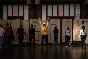 211106_MariaJoseWSGroup1_049 Some ten ZCMP participants face the back wall towards a row of colorful life-sized body maps. With backs towards the camera, some participants stand legs shoulder-length apart and arms raised, others jog in place, while some stare in concentration.