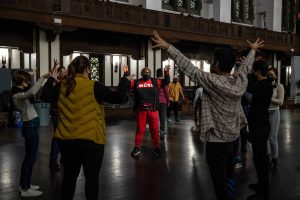 211106_MariaJoseWSGroup1_055 Two groups of ZCMP participants in a dimly lit church space engage in body movement exercises. In the foreground, the participants stand legs shoulder width apart, arms and fingers outstretched in a unified gesture.