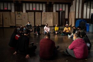 211106_MariaJoseWSGroup1_058 A circle of ZCMP participants speak and sit cross-legged on a dimly lit church space. In the back wall hang a row of colorful life-size body maps drawn on brown paper.