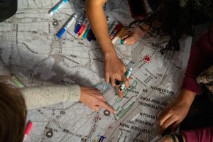 211106_MariaJoseWSGroup1_061 A black-and-white map of New York City is being drown upon by participants with an assortment of color markers, drawing shining red hearts, green paths in parks, bridges, and other placemarkers. Some hands point while others trace.