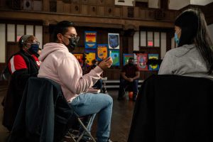 211106_MariaJoseWSGroup1_076 A close-up of two participants looking at each other as the one on the left, in jeans and pink hoodie, holds a lit match as they speak while the participant to the right with a gray sweater listens. Other participants in the semi-lit church space look on.