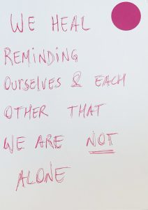Pink circle in the right hand corner. Pink text on a white background reads WE HEAL REMINDING OURSELVES AND EACH OTHER THAT WE ARE NOT ALONE