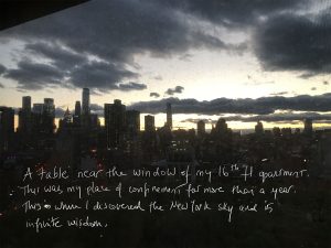 New York City skyline, with many tall buildings against a blue, gray and yellow sky. Several dark clouds in the sky. The bottom of the postcard features text which reads: A table near the window of my 16th fl apartment. This was my place of confinement for more than a year. This is where I discovered the New York sky and its infinite wisdom.