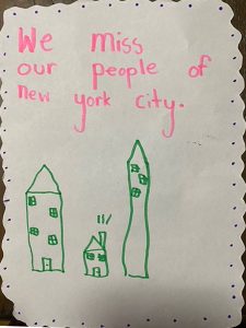 White postcard with three green drawings: one small house and two larger buildings. Pink text reads We miss our people of New York City.